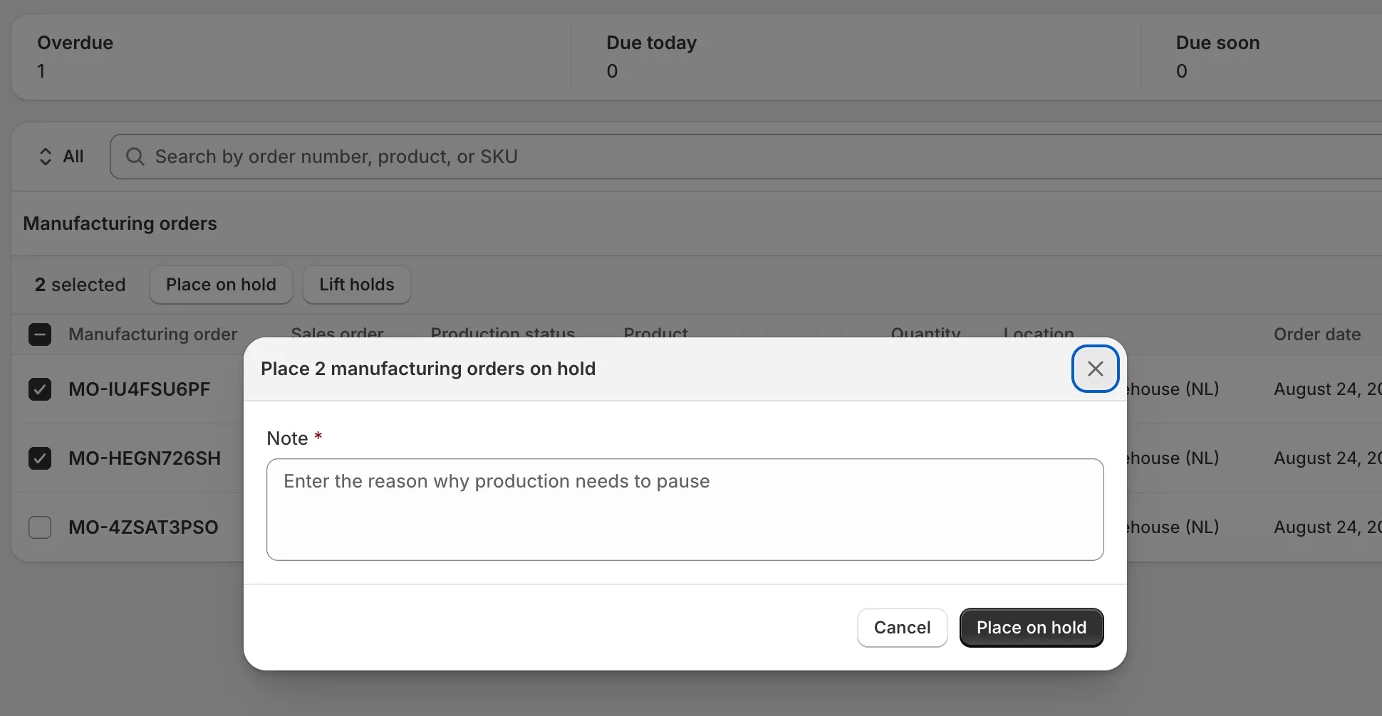 The manufacturing orders index with the bulk action bar above the list. Two rows are selected, and the bar reads “2 selected” with a Place on hold button open: a dialog asking for the reason why production needs to pause, with a required Note field, Cancel, and a Place on hold confirm.