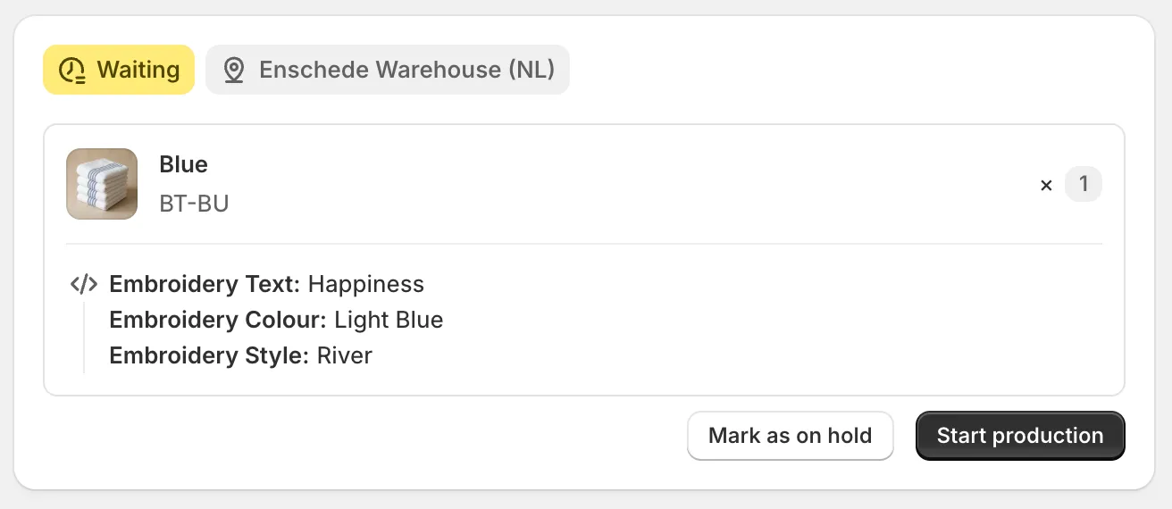 A manufacturing order in Orderkraft with a yellow Waiting badge and the Enschede Warehouse (NL) production location. Below it, one line for the Blue towel, SKU BT-BU, quantity one, with three customisations underneath: Embroidery Text is Happiness, Embroidery Colour is Light Blue, Embroidery Style is River. Mark as on hold and Start production buttons sit at the bottom.